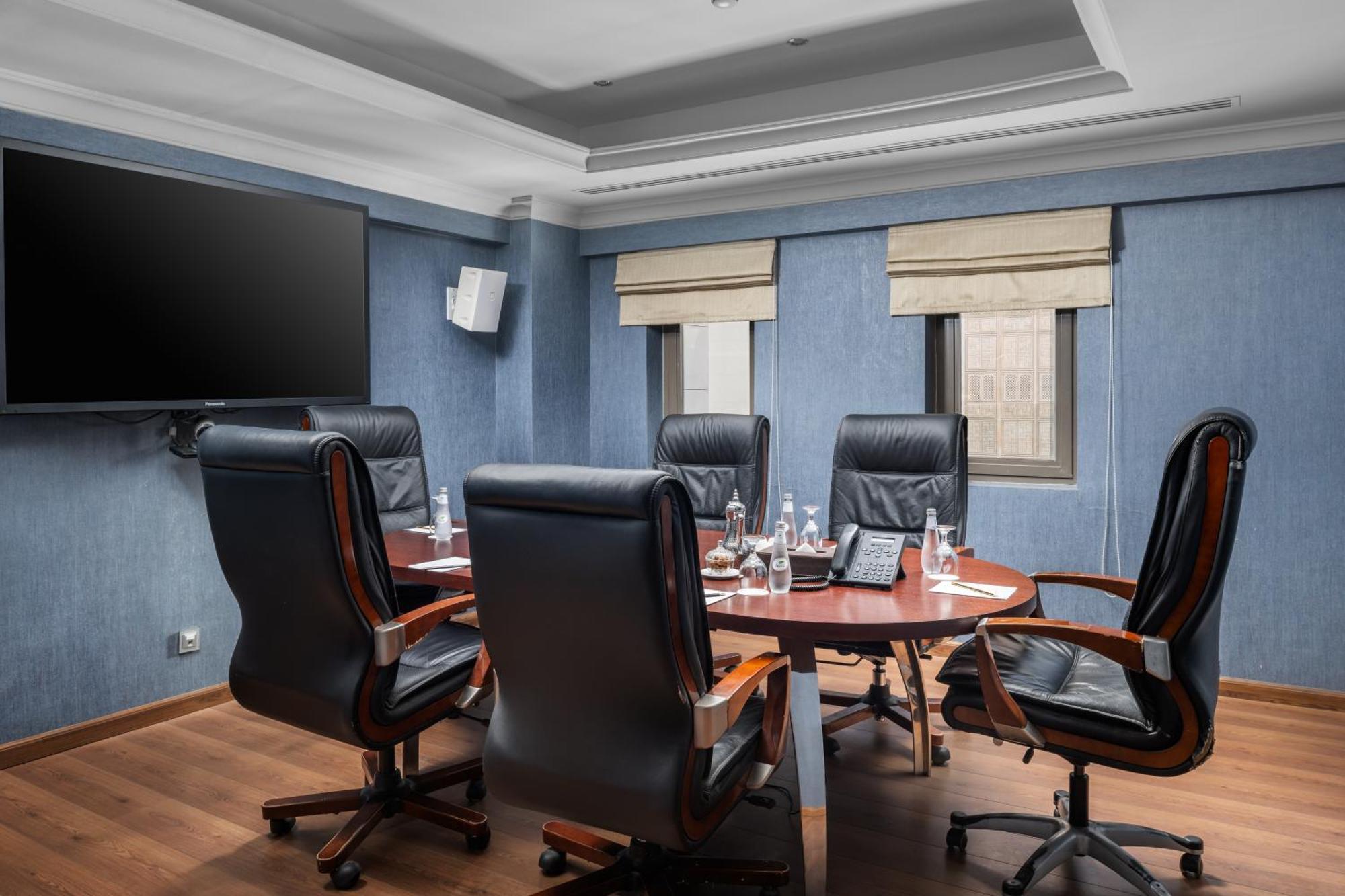 Al Aqeeq Madinah Hotel マディーナ エクステリア 写真 The photo shows a modern conference room. It features a round wooden table surrounded by several black leather swivel chairs. The walls have a blue color scheme, and there is a large flat-screen TV mounted on one wall. The room has two windows with b