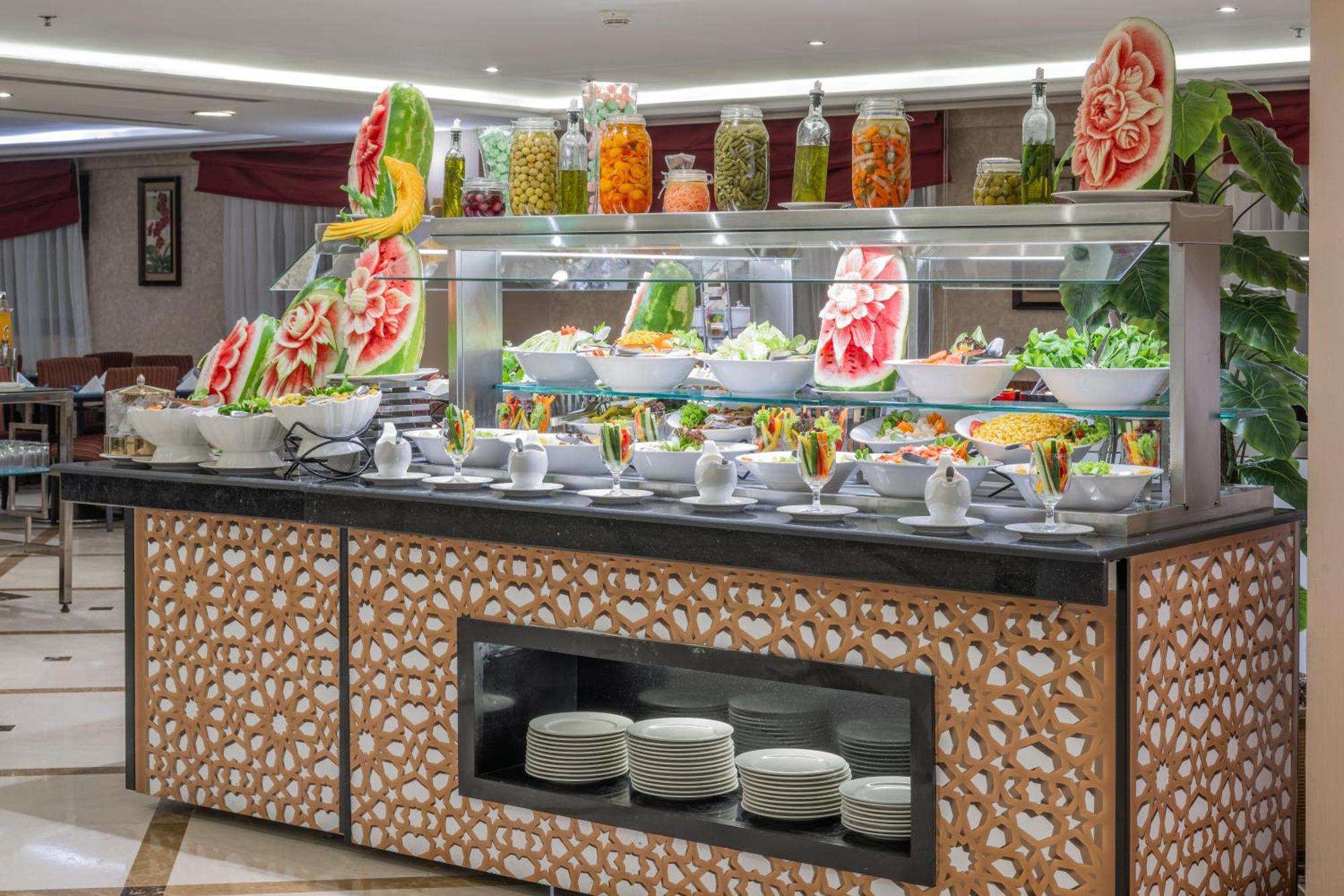 Al Aqeeq Madinah Hotel マディーナ エクステリア 写真 The photo shows a buffet display that features an array of colorful food items. In the foreground, there are several large bowls filled with various salads and dishes. The presentation is visually appealing, with decorative watermelon carvings and an