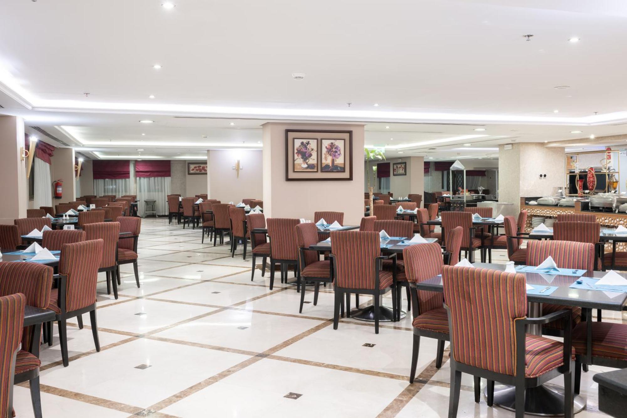 Al Aqeeq Madinah Hotel マディーナ エクステリア 写真 The photo shows the interior of a spacious restaurant. It features a well-organized dining area with neatly arranged tables and chairs. The tables are dressed with blue napkins, and there is a light-colored flooring with a grid pattern. The walls are