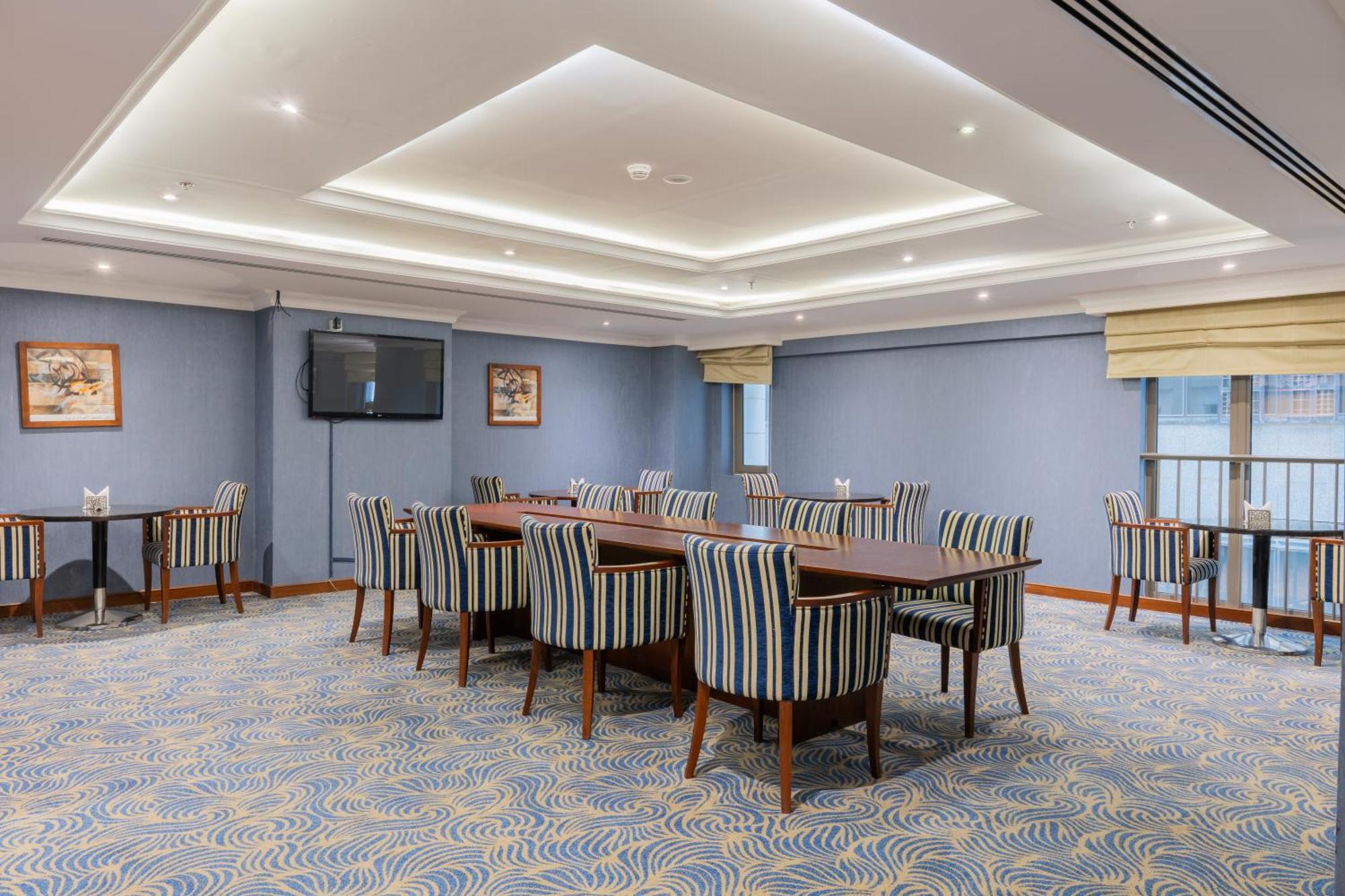 Al Aqeeq Madinah Hotel マディーナ エクステリア 写真 The photo shows a spacious conference or meeting room. It features a light blue color scheme with a patterned carpet. There are several rectangular tables arranged in a group, surrounded by striped chairs. The room is well-lit with modern ceiling lig