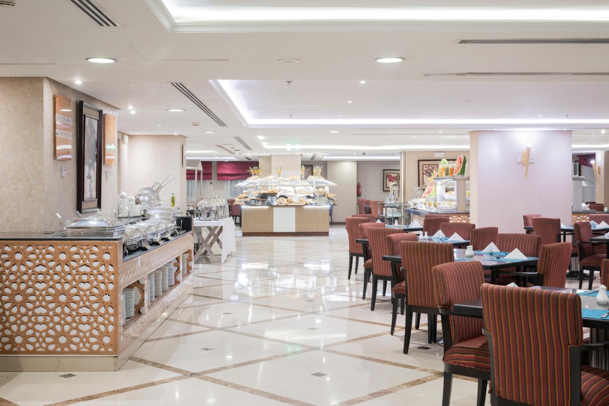 Al Aqeeq Madinah Hotel マディーナ エクステリア 写真 The photo shows an elegantly designed restaurant or dining area. It features a spacious layout with a glossy tiled floor and bright, soft lighting. There are several dining tables with striped chairs arranged neatly. Along one side, there's a buffet 