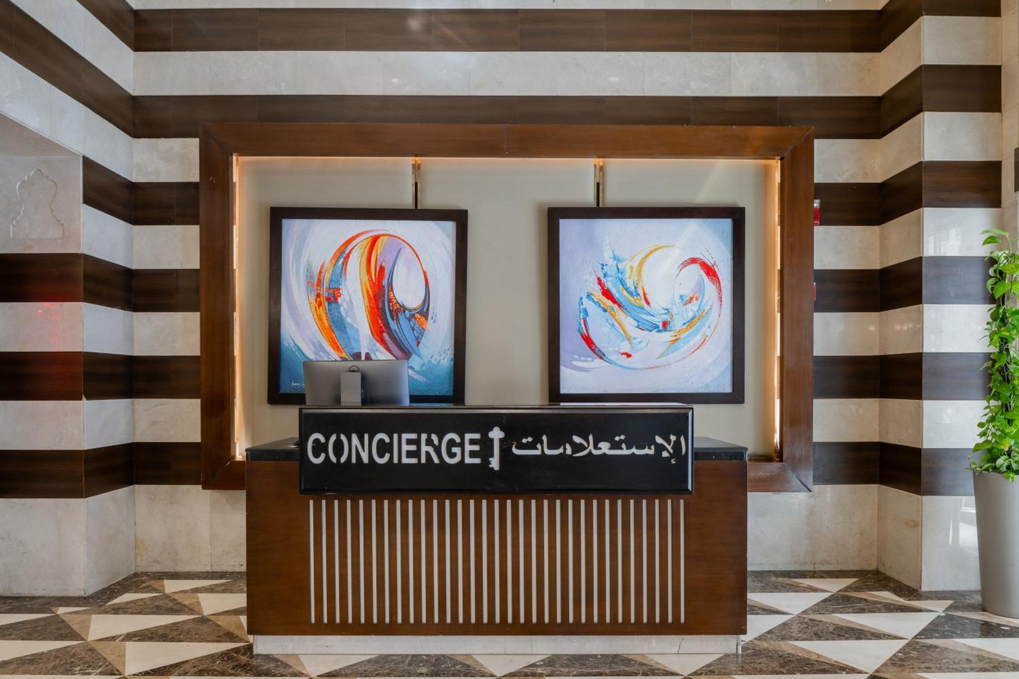 Al Aqeeq Madinah Hotel マディーナ エクステリア 写真 The image shows a concierge desk in a hotel lobby. The desk is made of wood with a striped design and features two large, colorful abstract paintings on the wall behind it. The sign on the desk reads "CONCIERGE" in English and has Arabic text below i