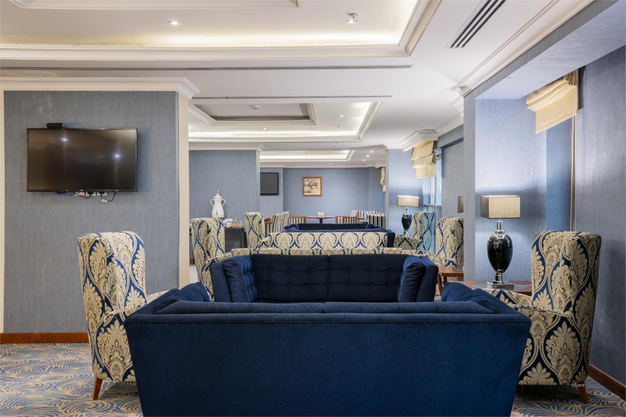 Al Aqeeq Madinah Hotel マディーナ エクステリア 写真 The photo shows a stylish and modern lounge area. There are two patterned armchairs with blue and cream designs, positioned next to a deep blue couch. The room has a light blue wall with a flat-screen TV mounted on one side. Soft lighting from stylis