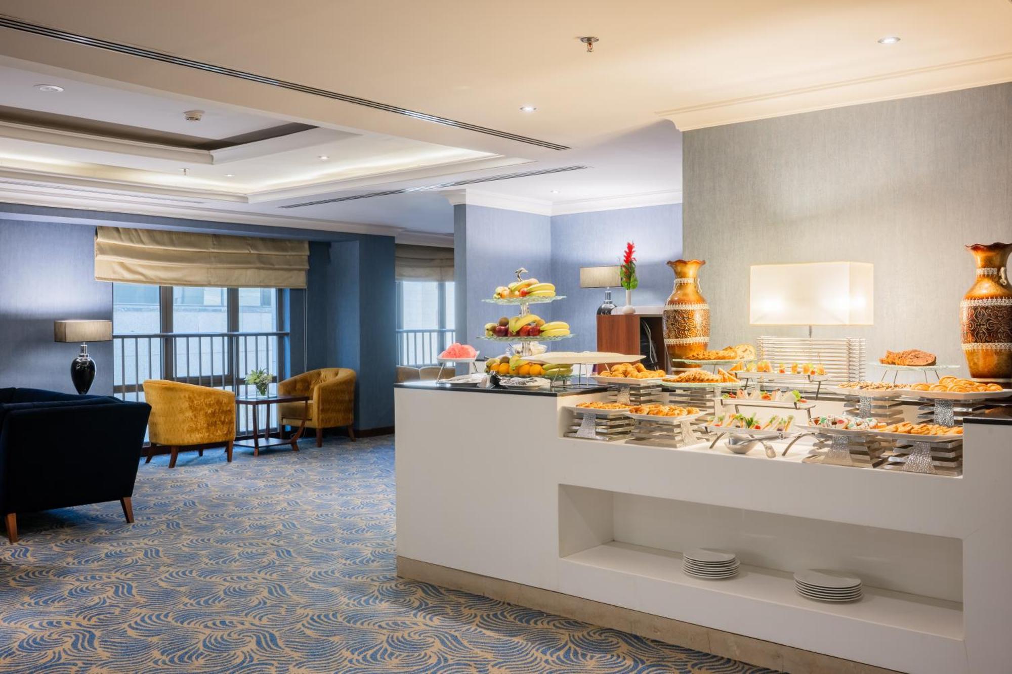 Al Aqeeq Madinah Hotel マディーナ エクステリア 写真 The photo shows a modern dining area, likely in a hotel or restaurant. On the right side, there is a buffet table with various food items displayed, including pastries and desserts. Behind the table, a decorative fountain is visible. The room feature