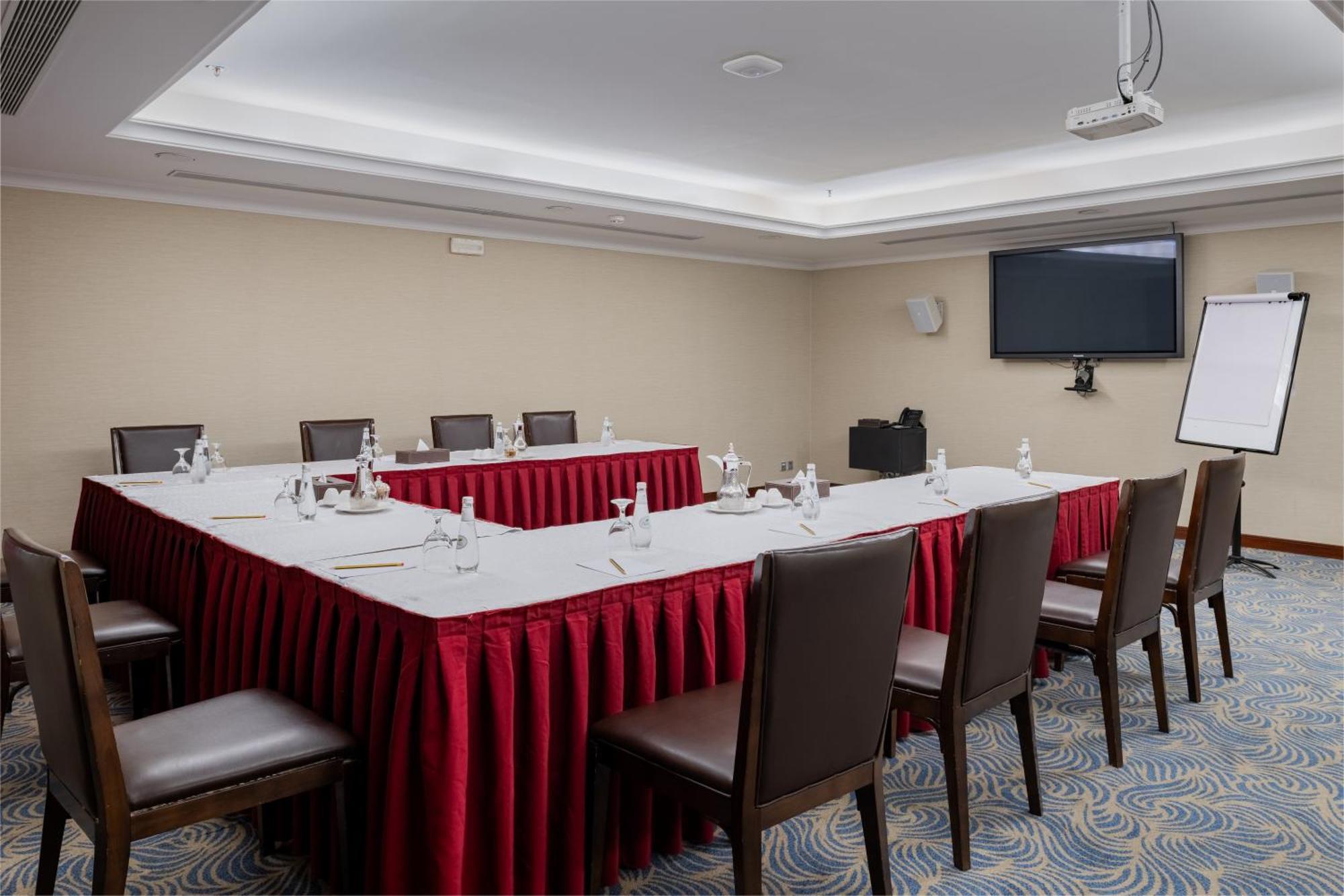 Al Aqeeq Madinah Hotel マディーナ エクステリア 写真 The image shows a conference room set up for a meeting. There is a U-shaped arrangement of tables covered with white linens and red table skirts. Each table position is equipped with a water bottle and a small notepad, suggesting that it is designed 