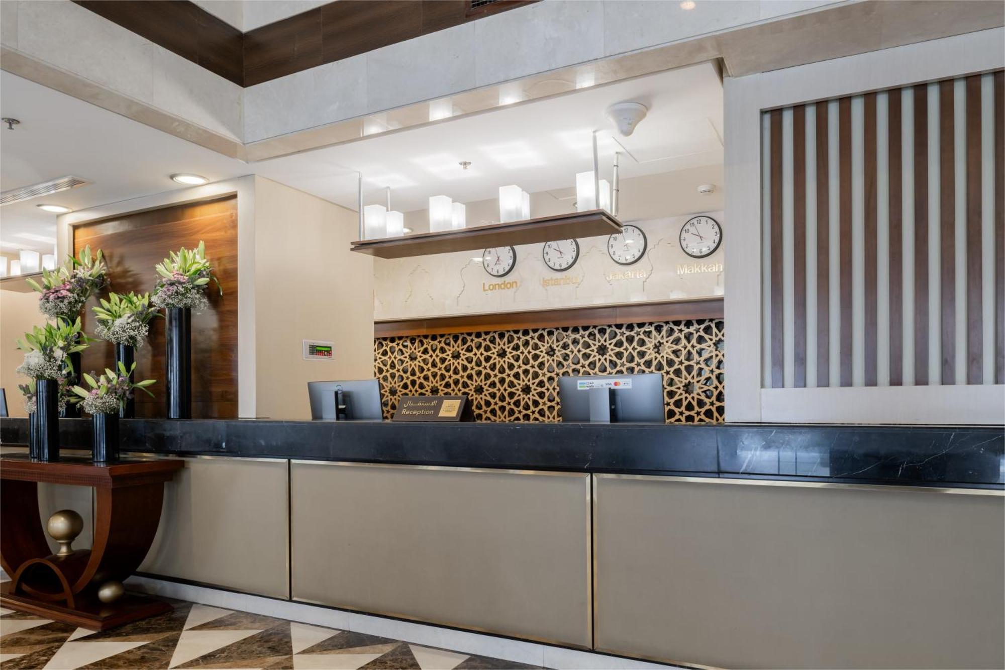 Al Aqeeq Madinah Hotel マディーナ エクステリア 写真 The photo shows the interior of a hotel reception area. The reception desk is designed with a sleek, modern look featuring a dark countertop and a light-colored front. Above the desk, there are several clocks displaying different times, suggesting an
