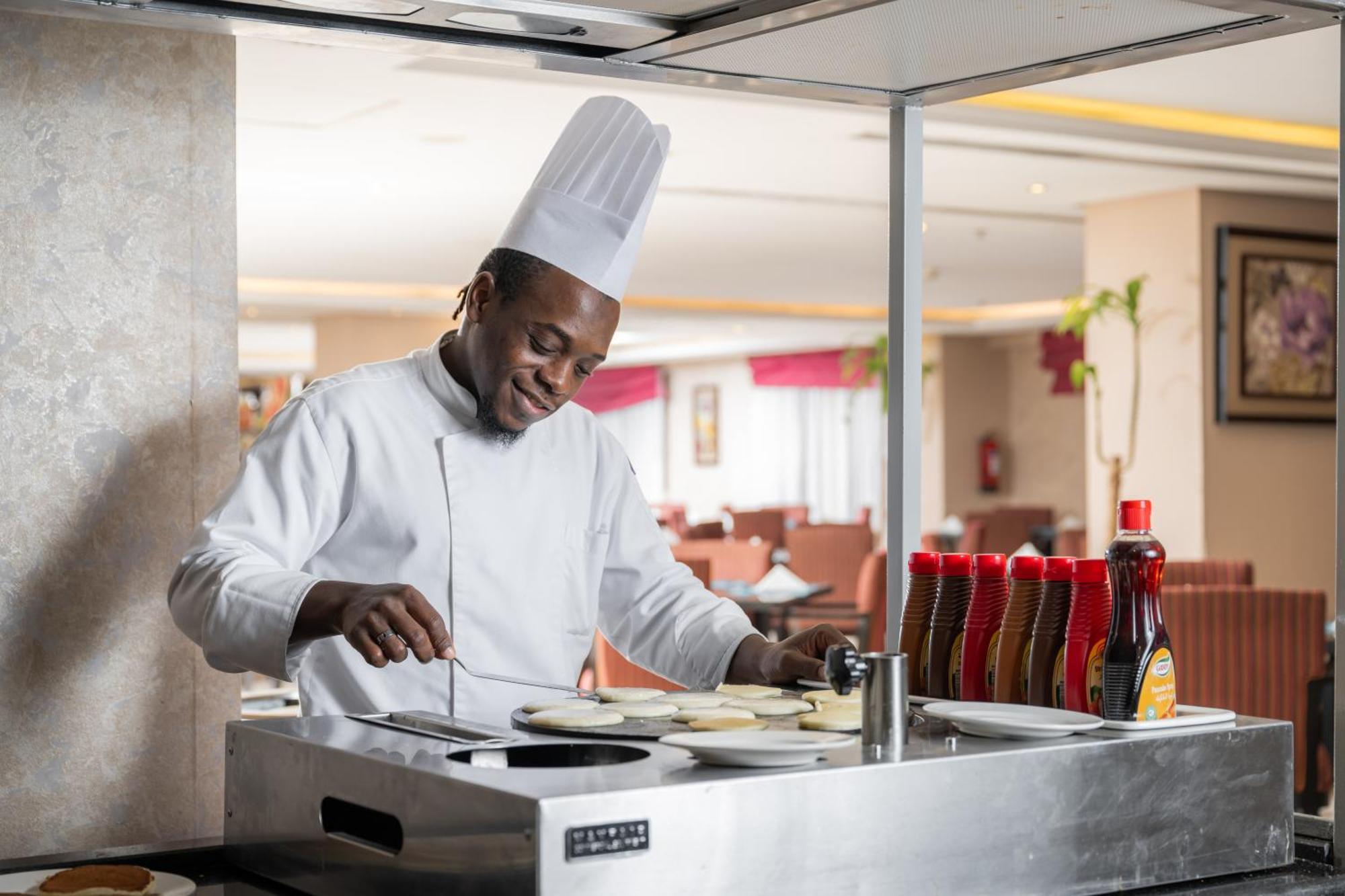 Al Aqeeq Madinah Hotel マディーナ エクステリア 写真 The photo shows a chef in a professional kitchen setting. He is wearing a traditional white chef's uniform and a tall white chef's hat, and he appears to be smiling while cooking. The chef is standing at a stainless steel cooking station, where he is