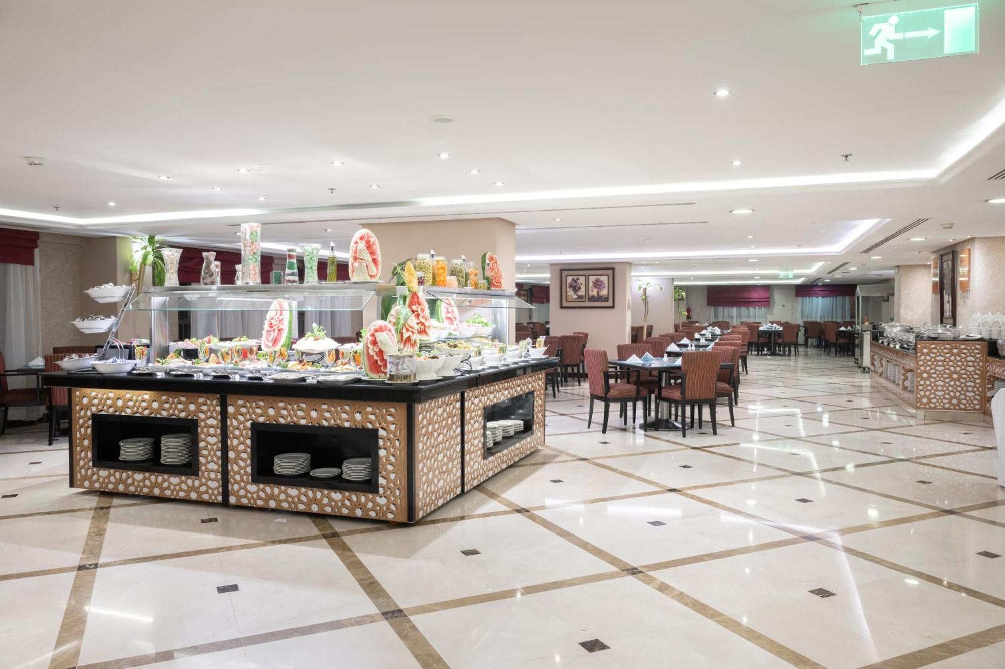 Al Aqeeq Madinah Hotel マディーナ エクステリア 写真 The photo shows a spacious restaurant interior with a modern design. In the foreground, there is a large buffet counter filled with various food items, beautifully arranged and decorated. The counter features a backlit display, showcasing fruits and 