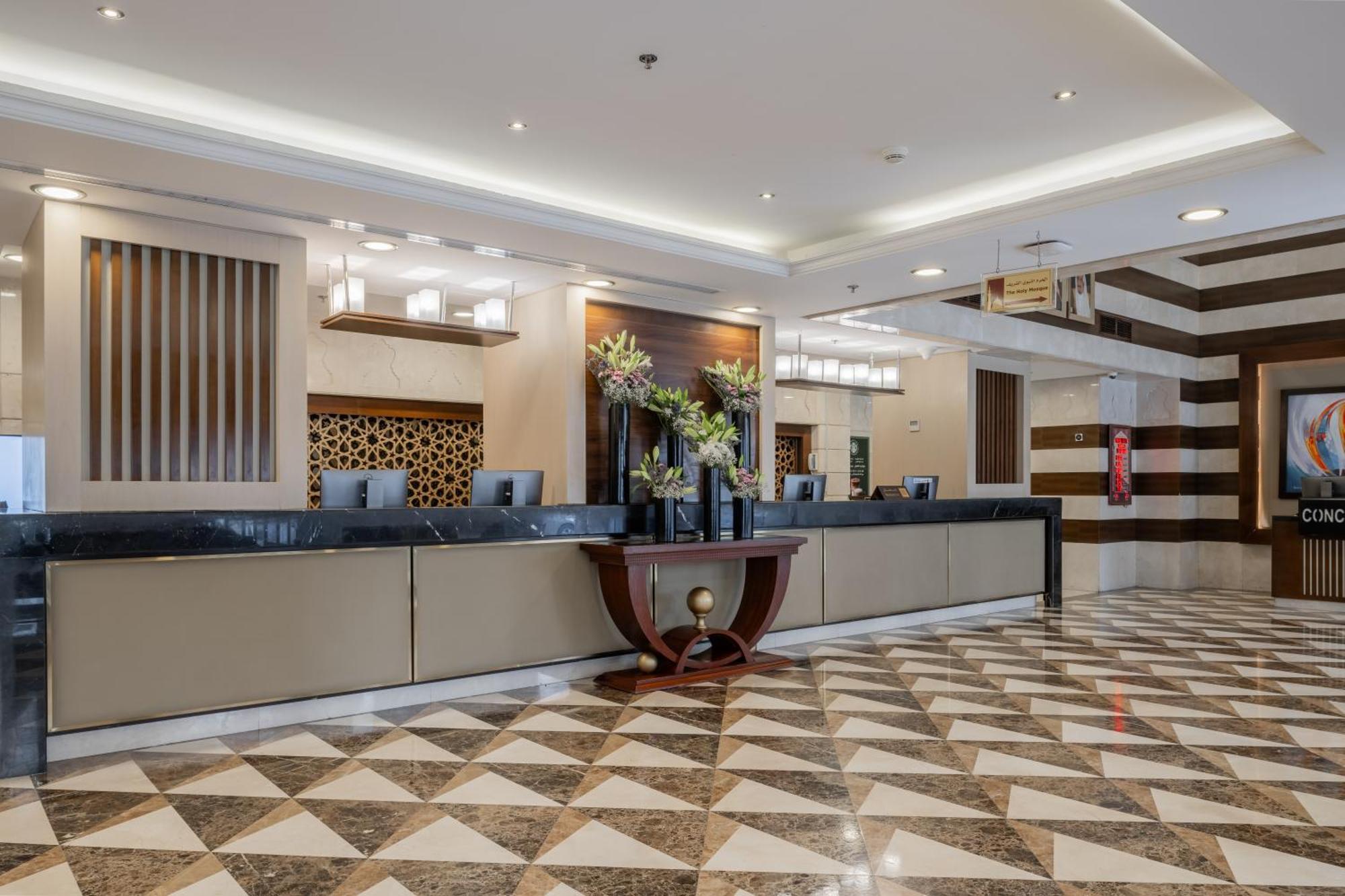 Al Aqeeq Madinah Hotel マディーナ エクステリア 写真 The photo depicts a hotel reception area. The check-in counter is long and sleek, with a smooth surface and decorative elements. Behind the counter, there are several computer monitors. The walls feature stylish designs, including wooden panels and g