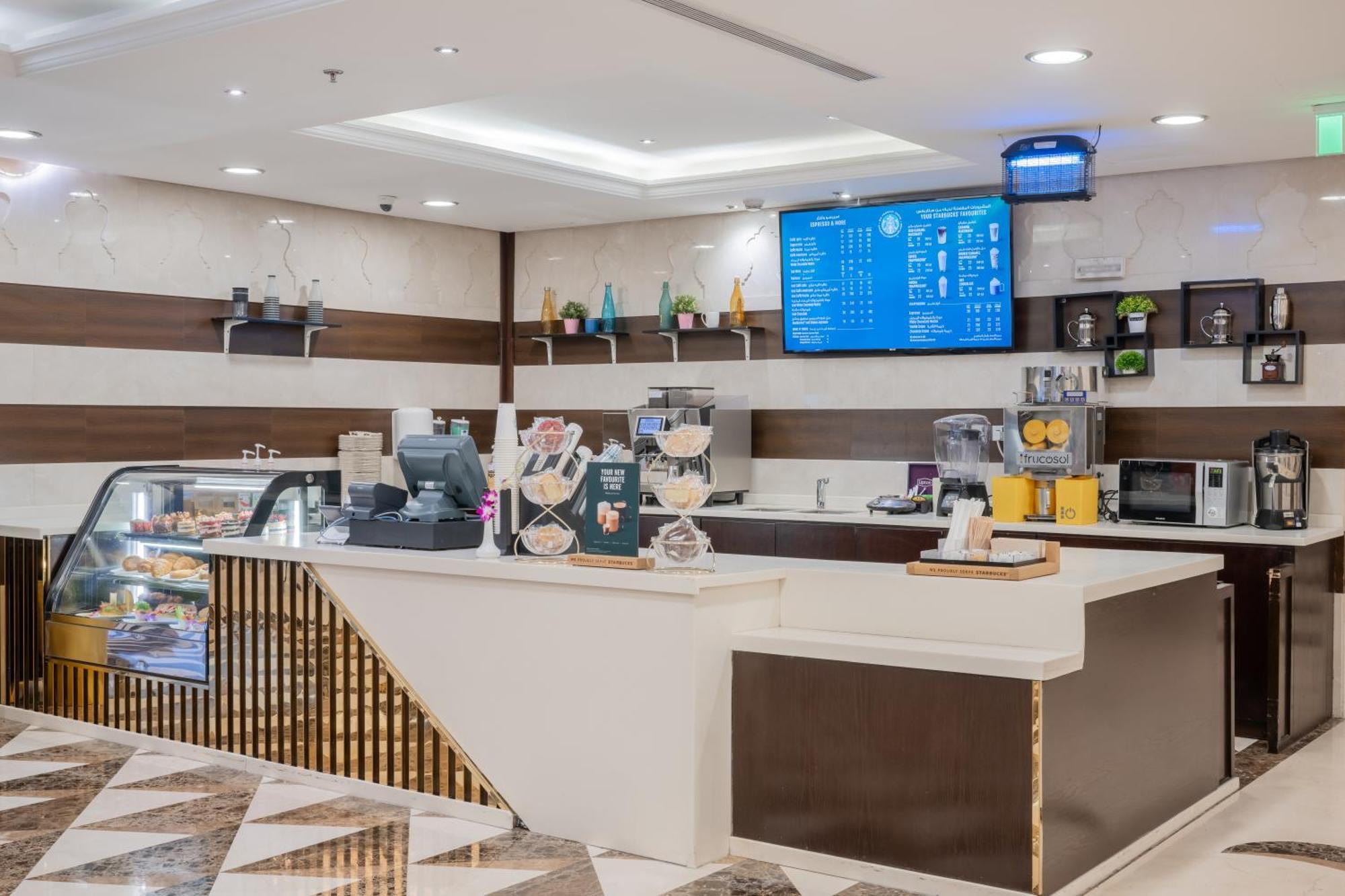 Al Aqeeq Madinah Hotel マディーナ エクステリア 写真 The photo shows a modern café or coffee shop interior. There's a counter area made of smooth white and dark brown materials, featuring a cash register and display items. Behind the counter, shelves are stocked with various beverage items and equipmen