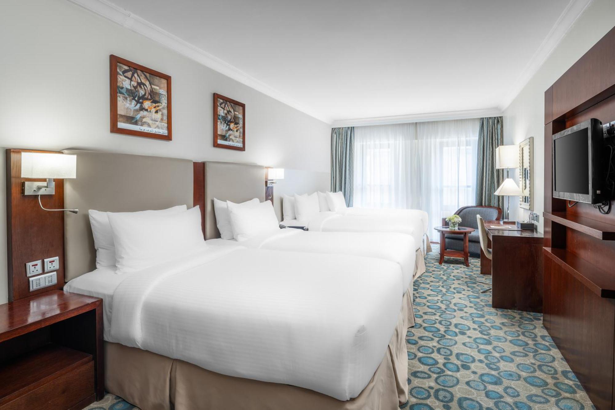 Al Aqeeq Madinah Hotel マディーナ エクステリア 写真 The photo shows a hotel room featuring a modern, clean design. There are two large beds with white linens positioned side by side. Above the beds are framed artworks, and soft lighting is provided by wall sconces. To the right, there is a wooden ente