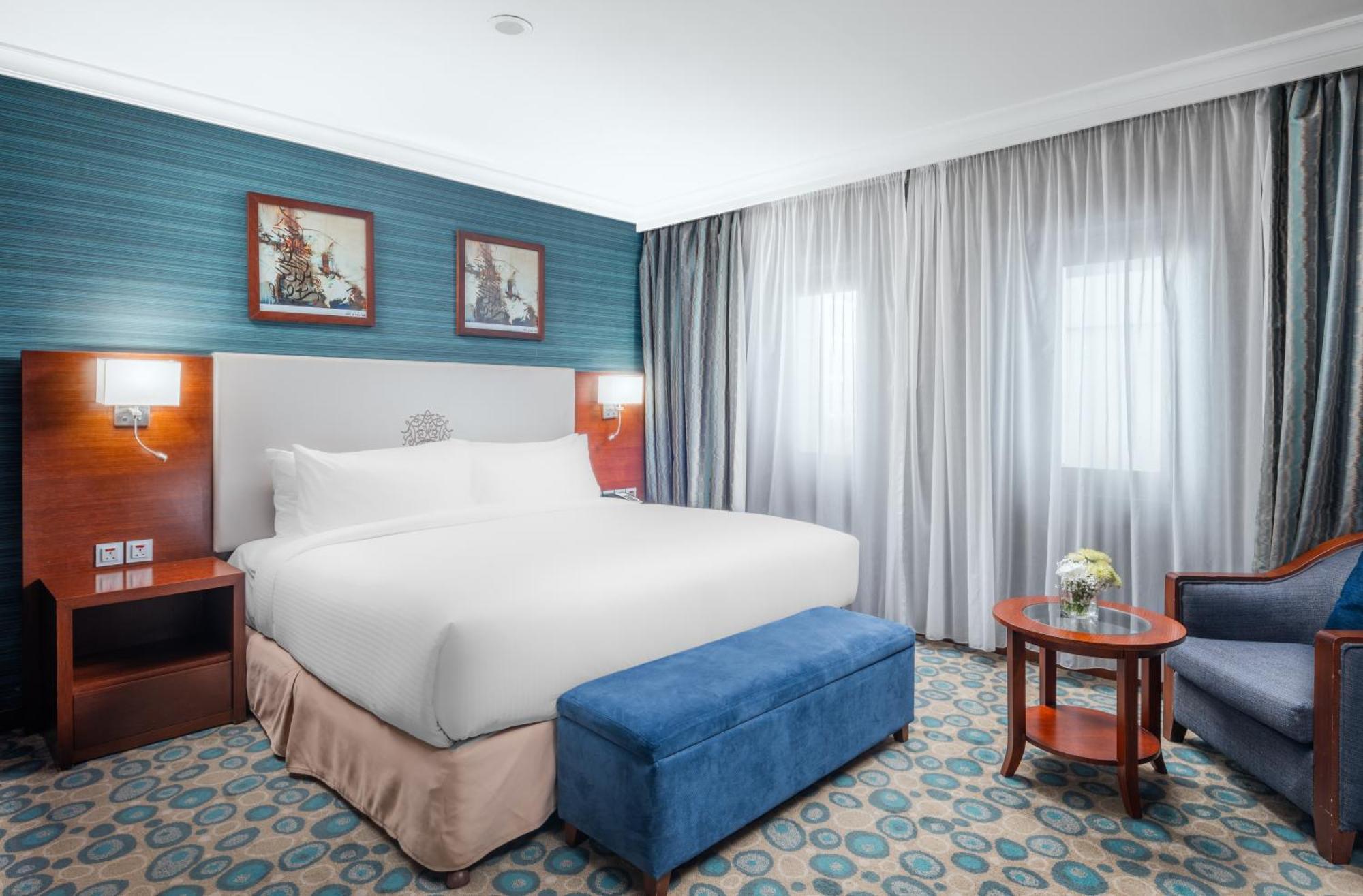 Al Aqeeq Madinah Hotel マディーナ エクステリア 写真 The photo shows a hotel room featuring a large, neatly made bed with a white duvet. The walls have a light blue accent with framed artwork above the bed. There’s a blue ottoman at the foot of the bed. To one side, there’s a small wooden table with a 
