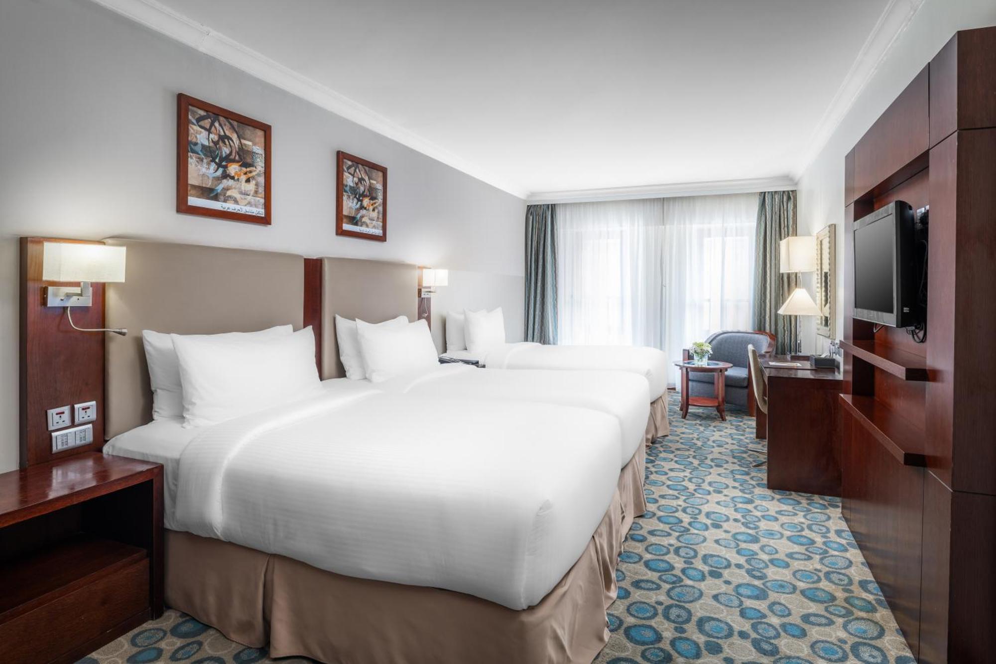 Al Aqeeq Madinah Hotel マディーナ エクステリア 写真 The photo shows a well-furnished hotel room featuring two large beds with white bedding. The walls are adorned with framed artwork. There are bedside lamps next to each bed, and a flat-screen TV is mounted on the wall. A large window allows natural l