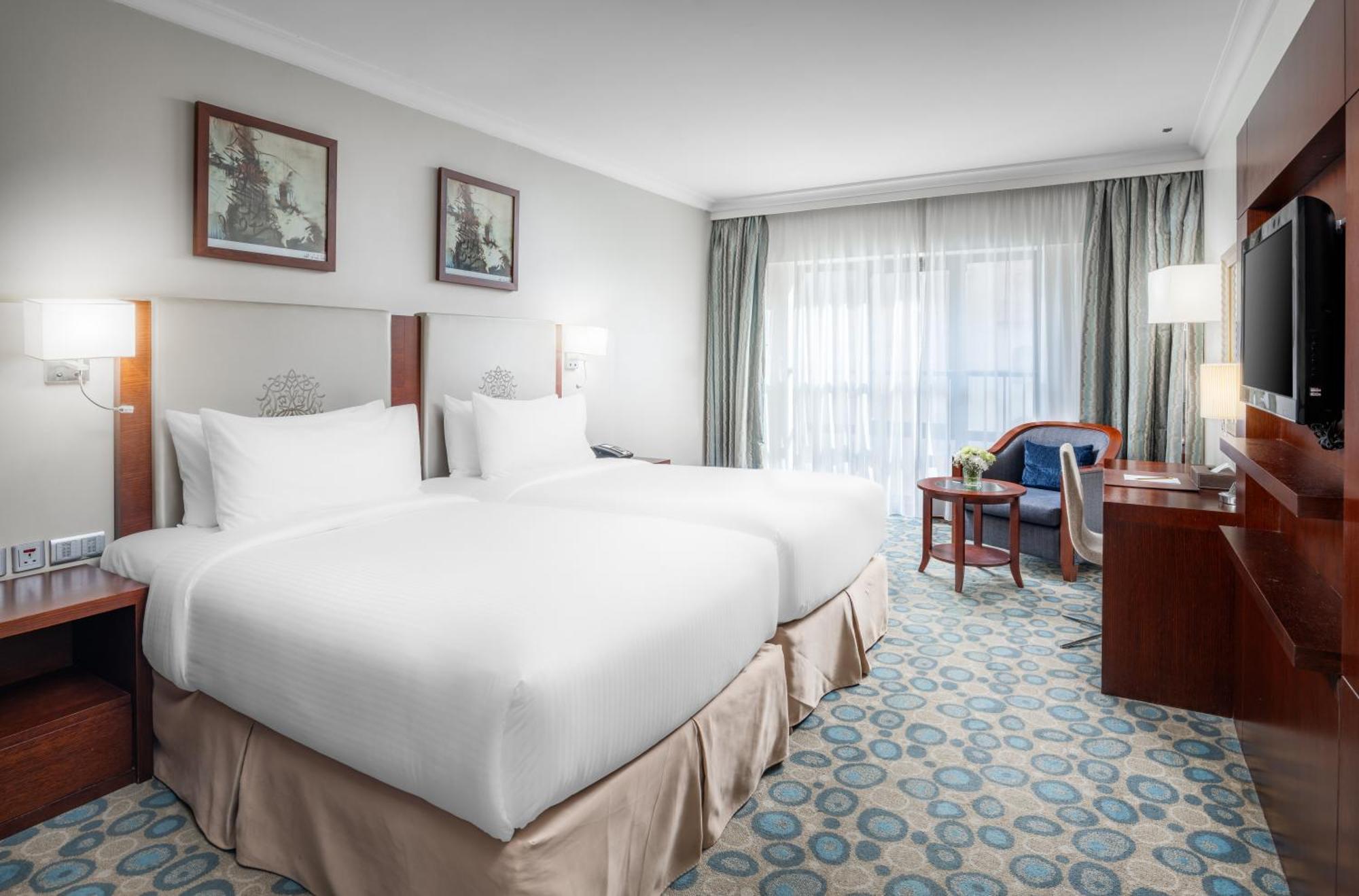 Al Aqeeq Madinah Hotel マディーナ エクステリア 写真 The photo shows a hotel room featuring two white beds with plush bedding. There are decorative artworks on the walls above the beds. A large window with sheer curtains allows natural light into the space, illuminating the room. The floor is covered w