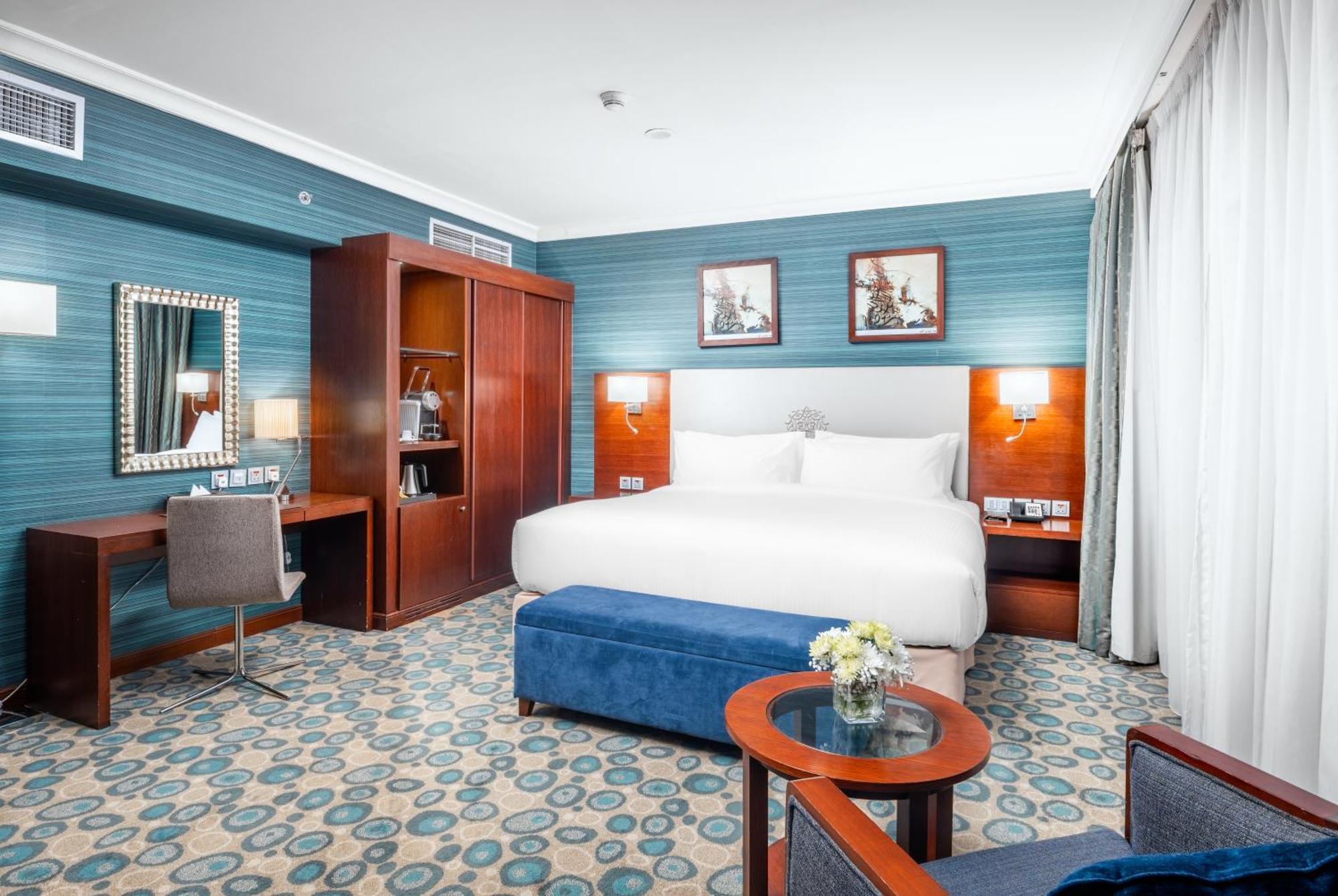 Al Aqeeq Madinah Hotel マディーナ エクステリア 写真 The photo shows a well-decorated hotel room. It features a large bed with white bedding, flanked by wall-mounted lights. Behind the bed, there are two framed pictures hanging on a blue wall. To the left, there's a wooden wardrobe and a desk with a ch
