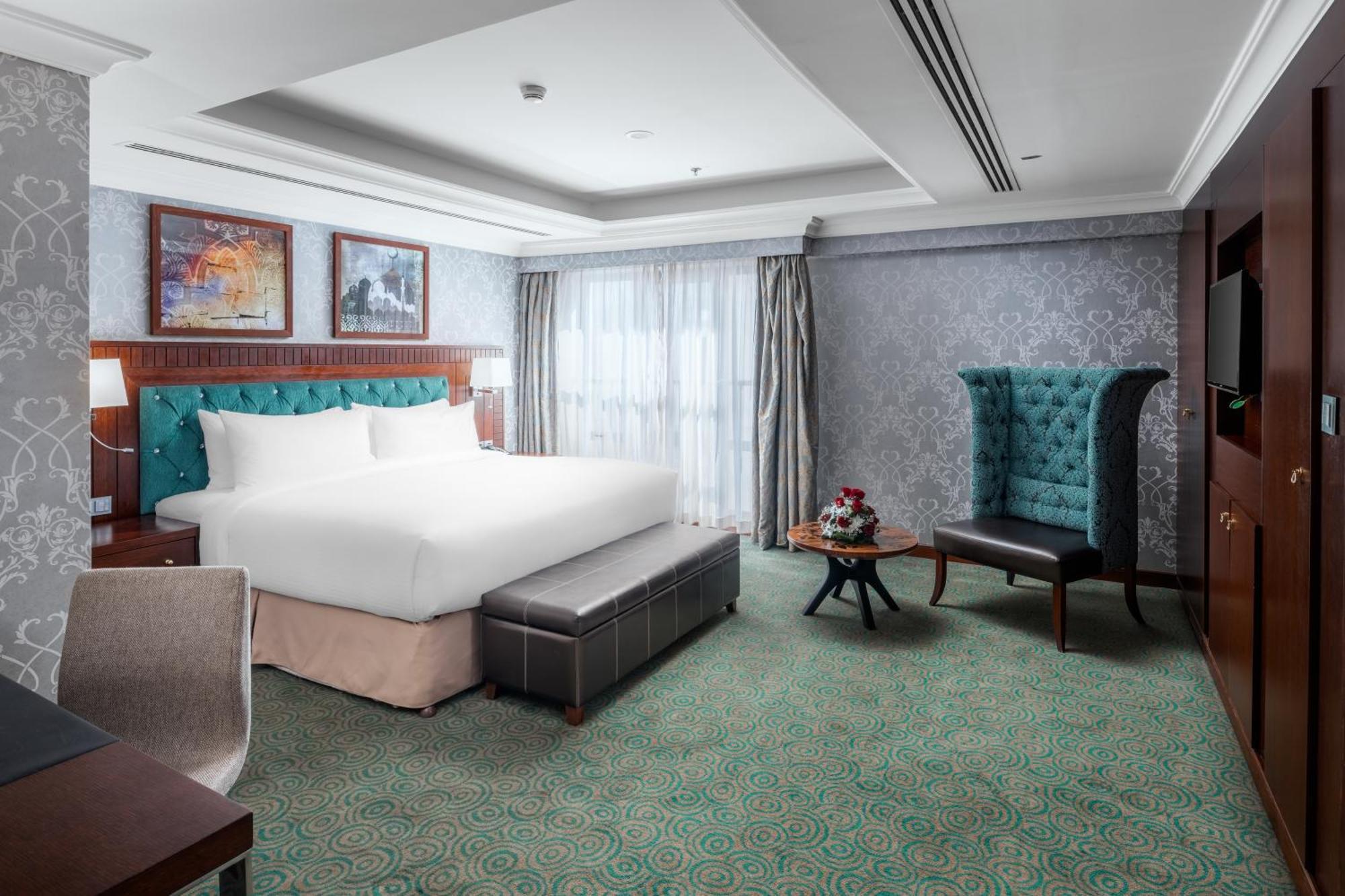 Al Aqeeq Madinah Hotel マディーナ エクステリア 写真 The photo shows a well-appointed hotel room. It features a large bed with a tufted headboard in a turquoise color, complemented by decorative pillows. There is a comfortable ottoman at the foot of the bed and a small coffee table with a vase of flowe