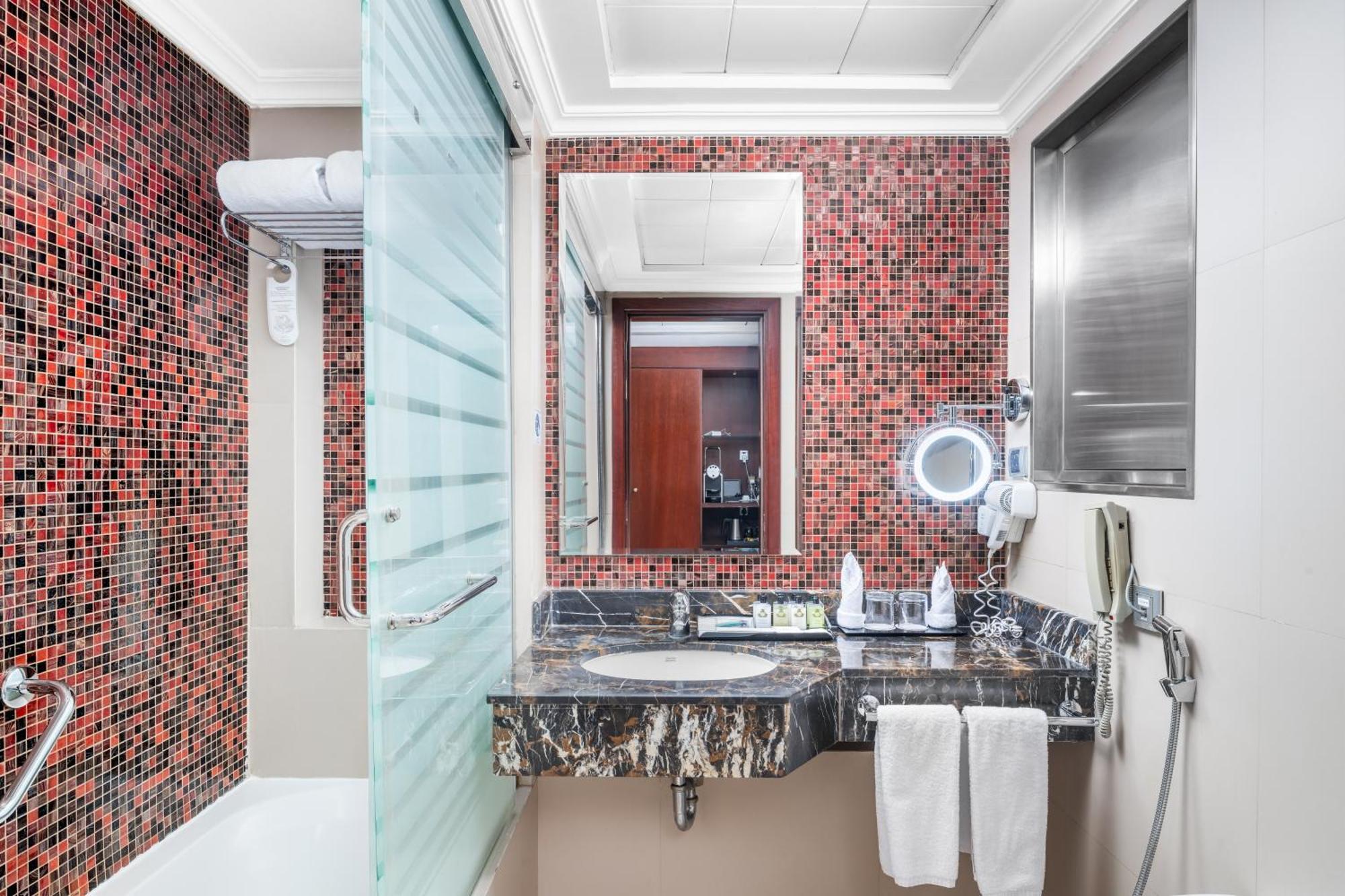 Al Aqeeq Madinah Hotel マディーナ エクステリア 写真 The photo shows a modern bathroom featuring a mix of design elements. The walls have a vibrant red mosaic tile pattern, adding a bold touch to the space. There is a glass shower enclosure, and next to it, a sleek countertop with a dark, marbled surfa