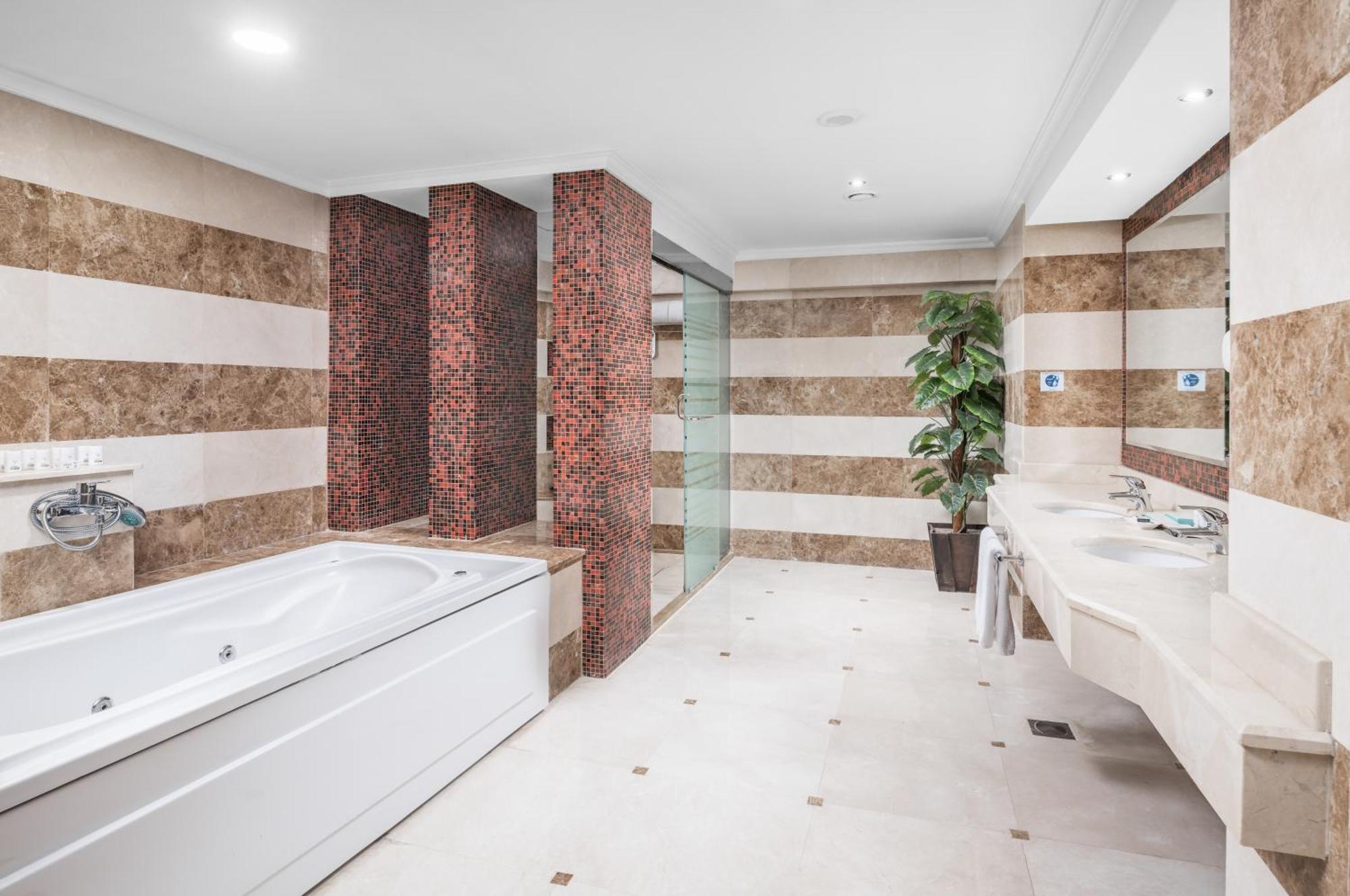 Al Aqeeq Madinah Hotel マディーナ エクステリア 写真 The photo shows a modern bathroom featuring a spacious layout. On one side, there is a large, white bathtub adjacent to a textured wall with warm, earthy tones. The walls are decorated with alternating horizontal stripes of light and dark colors. 

I
