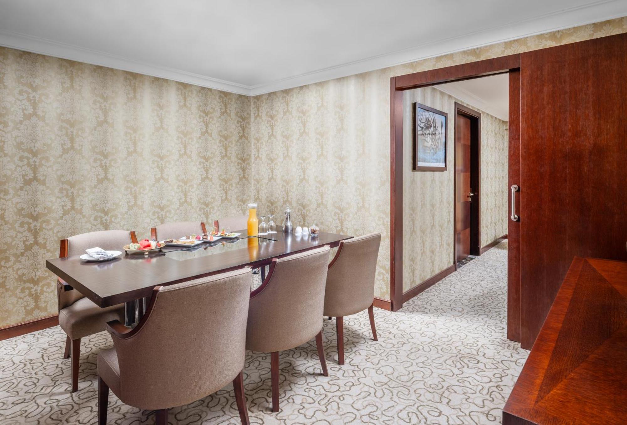 Al Aqeeq Madinah Hotel マディーナ エクステリア 写真 The photo shows a dining room with a stylish and elegant design. There is a large wooden dining table surrounded by plush, beige chairs. On the table, there are various items including a bottle of orange juice, glasses, and other dining accessories. 