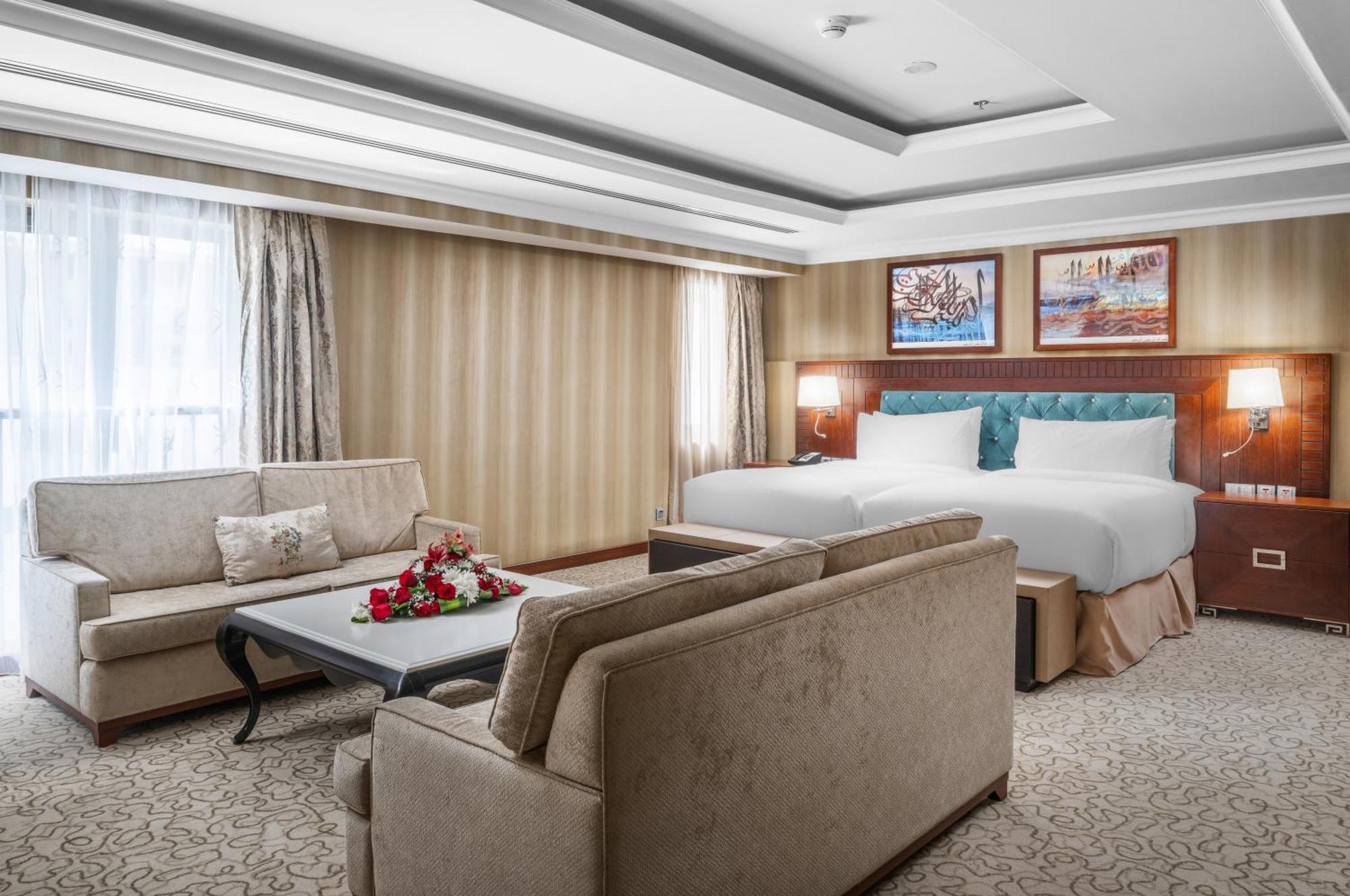 Al Aqeeq Madinah Hotel マディーナ エクステリア 写真 The photo shows a well-decorated hotel room. It features a large bed with two pillows and a light-colored duvet. There are two comfortable couches arranged around a coffee table with a floral centerpiece. The walls have a subtle striped design, and l