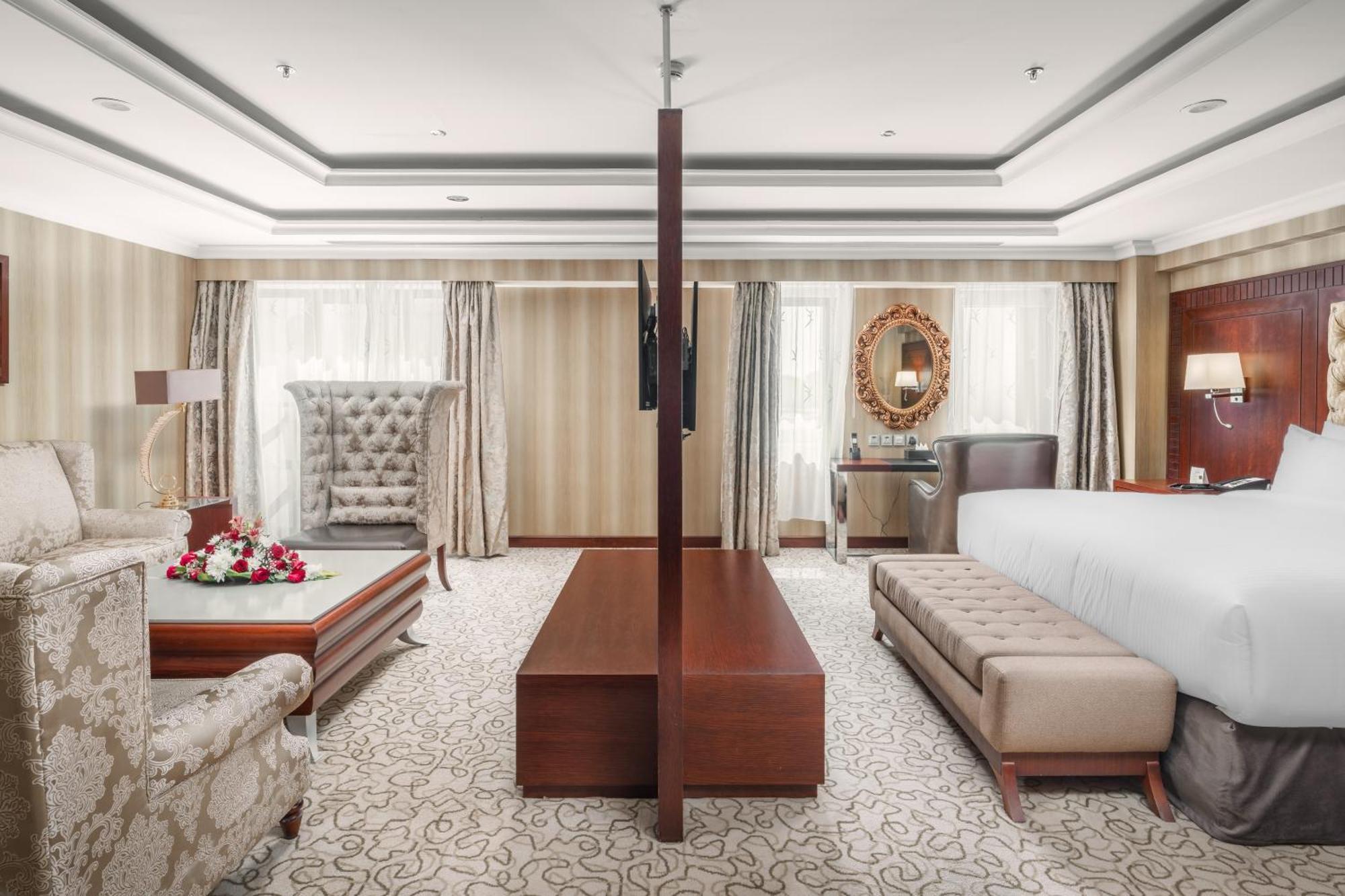 Al Aqeeq Madinah Hotel マディーナ エクステリア 写真 The photo showcases a spacious hotel suite with an elegant and modern design. The room features a large bed on one side, complemented by a plush bench at the foot. Opposite the bed, there is a stylish seating area that includes a decorative armchair 