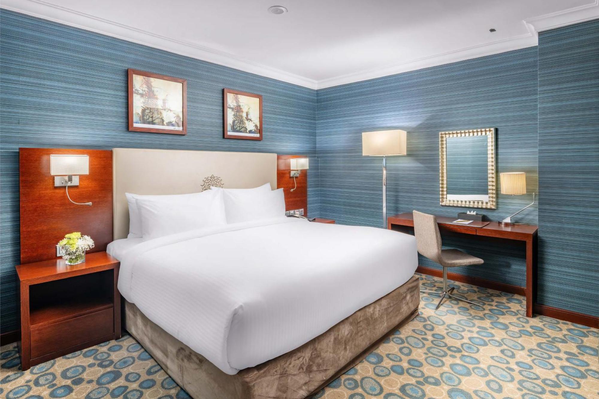 Al Aqeeq Madinah Hotel マディーナ エクステリア 写真 The photo shows a well-furnished hotel room. It features a large bed with white linens, flanked by wooden nightstands. The walls are painted in a light blue color with a textured pattern. Above the bed, there are two framed pictures. On one side of t