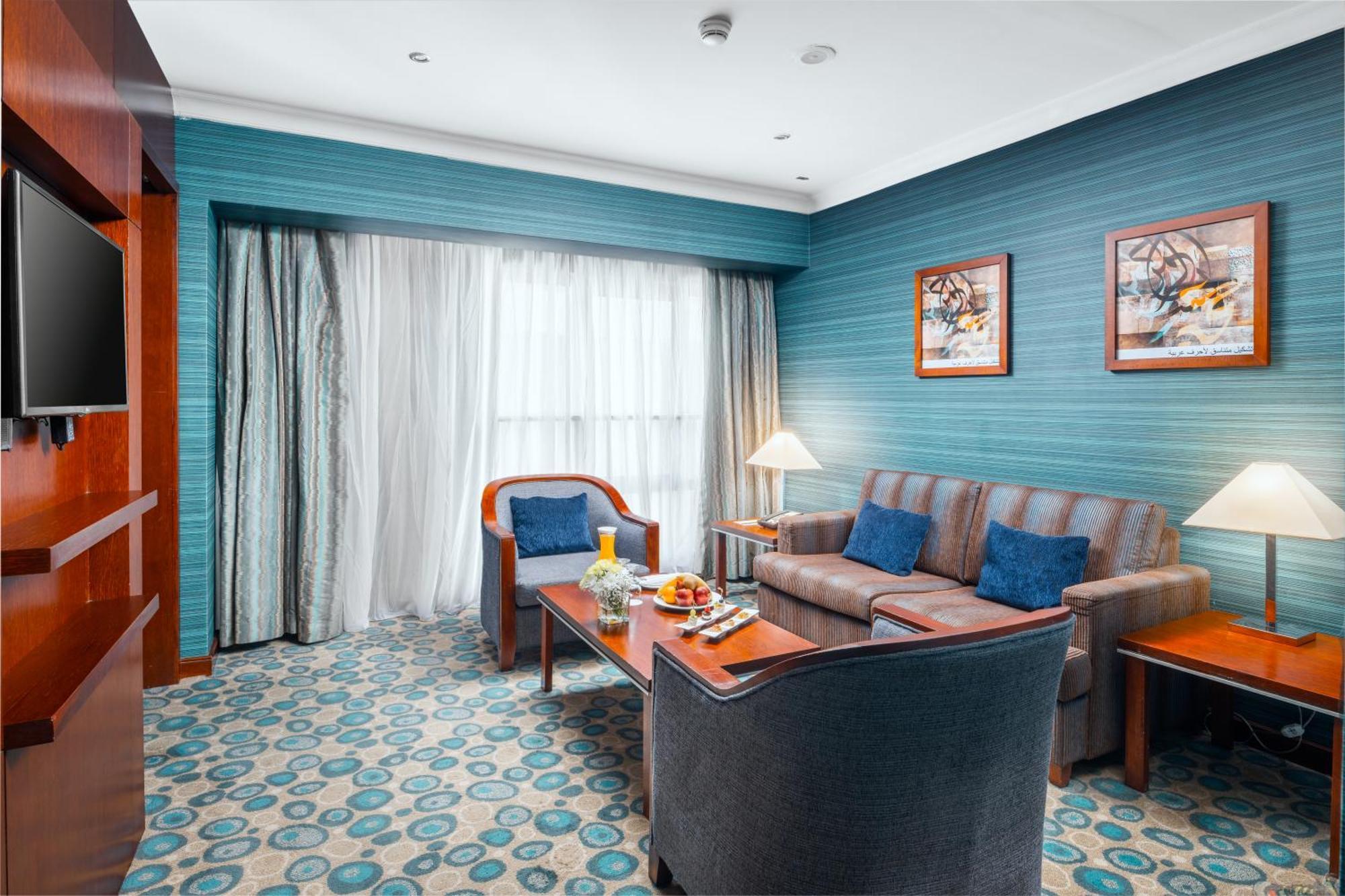 Al Aqeeq Madinah Hotel マディーナ エクステリア 写真 The photo shows a cozy and modern hotel room featuring a sitting area. The walls are painted a calming teal color, complemented by a patterned carpet in shades of blue and beige. There is a comfortable sofa with cushions, two armchairs, and a wooden 