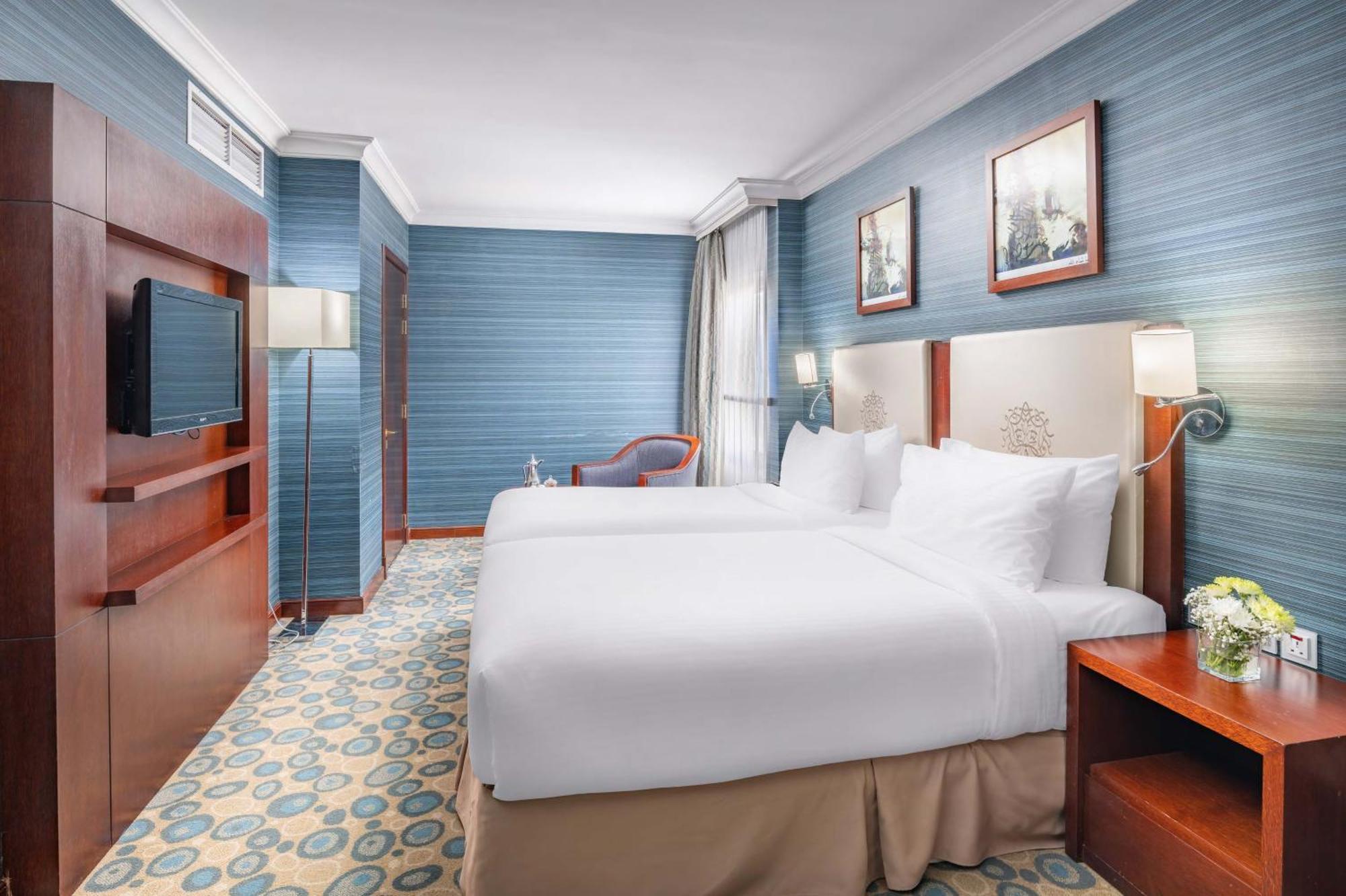 Al Aqeeq Madinah Hotel マディーナ エクステリア 写真 The photo shows a hotel room featuring two twin beds dressed in white linens. The walls are painted in a soft blue color, and there are framed artworks hung above the beds. A flat-screen TV is mounted on a wooden unit against one wall. To the side, t