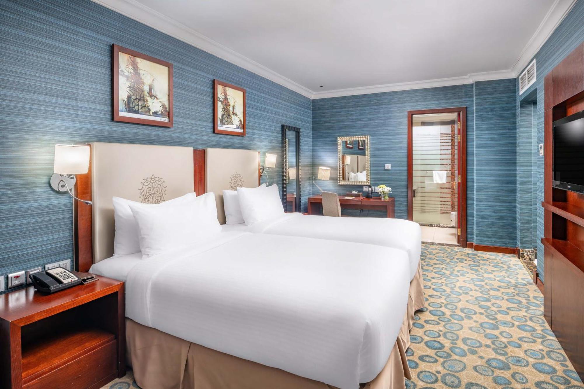 Al Aqeeq Madinah Hotel マディーナ エクステリア 写真 The photo shows a hotel room featuring two twin beds with white bedding, positioned against a light-colored headboard. The walls are painted in a soft blue shade, adorned with framed artwork. There is a wooden desk with a chair, and a mirror mounted 