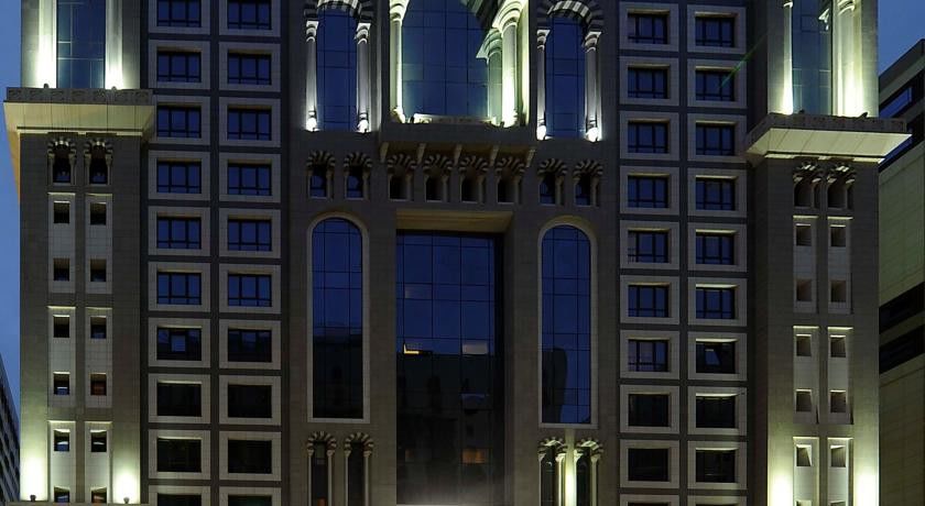 Al Aqeeq Madinah Hotel マディーナ エクステリア 写真 The image shows the facade of a modern high-rise building illuminated at night. The design features large glass windows arranged in a grid pattern, along with decorative architectural elements at the top, including arches and ornate detailing. The co
