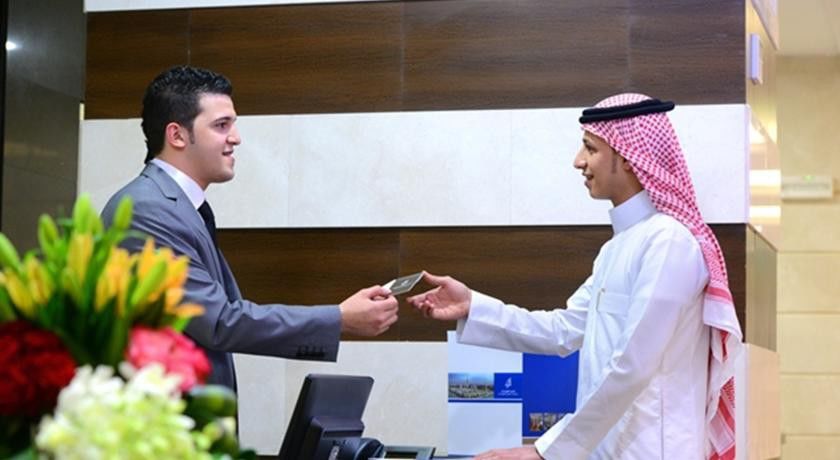 Al Aqeeq Madinah Hotel マディーナ エクステリア 写真 The image depicts two individuals at a reception desk. The person on the left, dressed in a formal suit, appears to be handing a card or document to the person on the right, who is wearing traditional attire, including a white thobe and a red and whi