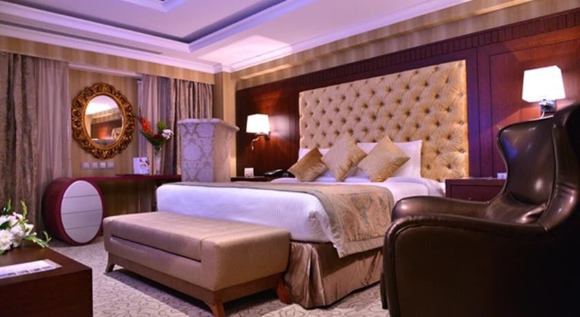 Al Aqeeq Madinah Hotel マディーナ エクステリア 写真 The photo shows a luxurious hotel room. It features a large, upholstered headboard with a tufted design, paired with plush bedding and decorative cushions. There's a stylish bench at the foot of the bed and a comfortable armchair to the side. The roo