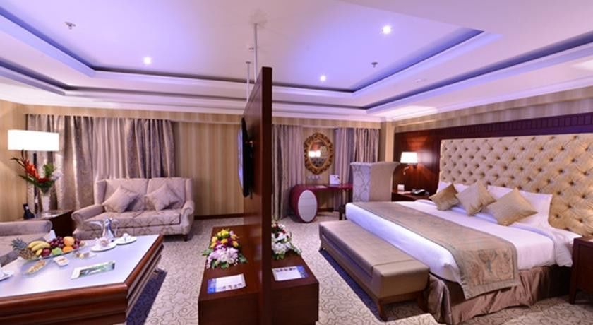 Al Aqeeq Madinah Hotel マディーナ エクステリア 写真 The image shows a luxurious hotel room. It features a large bed with a tufted headboard, flanked by nightstands with lamps. There's a seating area with a plush sofa and a coffee table. A decorative partition separates the bed from the living area. Th