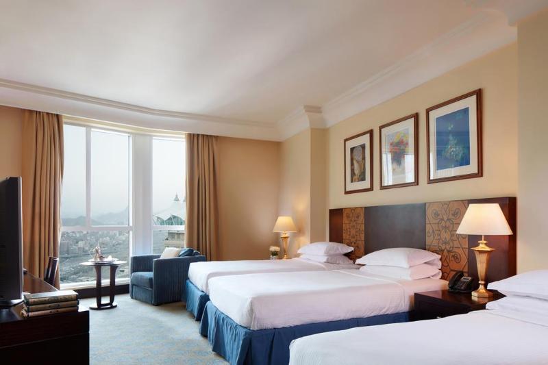 Al Aqeeq Madinah Hotel マディーナ エクステリア 写真 The photo shows a hotel room featuring three beds arranged neatly. The room has a warm, inviting ambiance with beige walls and a soft carpet. Large windows allow natural light to fill the space, offering a view outside. There are curtains in a light 