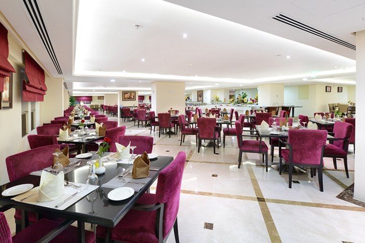 Al Aqeeq Madinah Hotel マディーナ エクステリア 写真 The photo shows an elegant restaurant interior with a spacious layout. The dining area features multiple tables, each set with white tablecloths and dinnerware. The chairs are upholstered in a rich purple fabric, adding a touch of luxury to the atmos