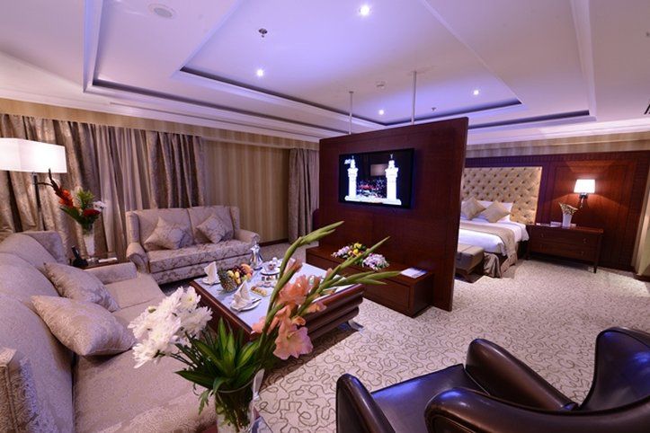 Al Aqeeq Madinah Hotel マディーナ エクステリア 写真 The photo shows a luxurious hotel room suite. There is a comfortable seating area with stylish sofas and a coffee table. A decorative arrangement of flowers is on the table, adding a touch of elegance. A large flat-screen television is mounted on a p
