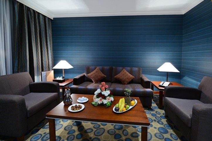 Al Aqeeq Madinah Hotel マディーナ エクステリア 写真 The photo shows a cozy living area featuring a dark blue wall with a textured design. In the center, there is a wooden coffee table surrounded by two comfortable armchairs and a sofa with decorative cushions. On the table, there are plates with snack