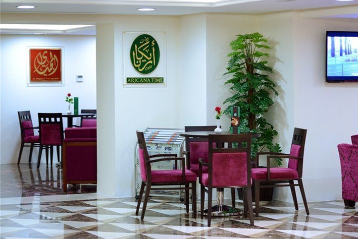 Al Aqeeq Madinah Hotel マディーナ エクステリア 写真 The photo shows an interior space, likely a dining or lounge area. There are several tables with four chairs each, featuring a modern design with dark wood and plush maroon upholstery. On the walls, there are decorative signs, one of which has Arabic