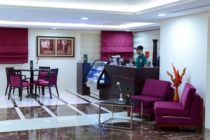 Al Aqeeq Madinah Hotel マディーナ エクステリア 写真 The photo shows a modern interior space, likely a café or small restaurant. It features a seating area with a few tables and chairs, all in a dark wood finish with purple upholstery. There is a counter area with a staff member behind it, possibly ser