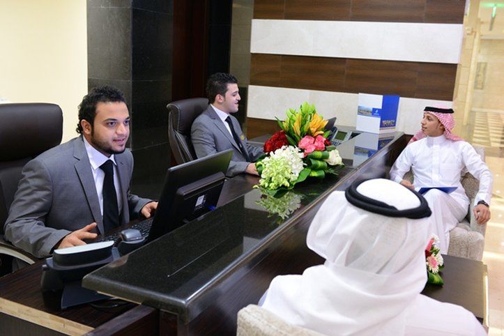 Al Aqeeq Madinah Hotel マディーナ エクステリア 写真 The photo depicts a reception area with three individuals. Two men are seated behind a reception desk; they are dressed in formal suits and appear to be engaged in conversation, possibly assisting a guest. The desk has a computer and a vibrant arrang