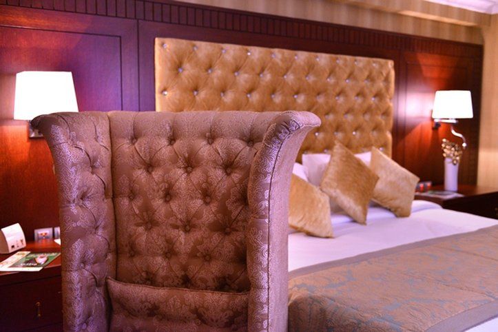 Al Aqeeq Madinah Hotel マディーナ エクステリア 写真 The image shows a luxurious bedroom featuring a large upholstered headboard with a tufted design in a soft, beige color. The bed is elegantly dressed with decorative pillows and a textured bedspread. In the background, there are wooden paneling and s